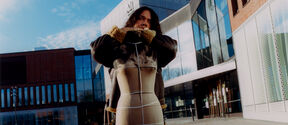 Person wearing a leather coat, stands in front of Aalto University's modern campus building with large windows.