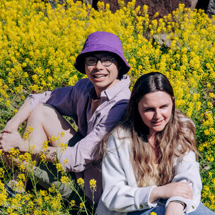 Two Aalto University students sitting outdoors in a field of yellow flowers, one wearing a purple hat and the other in a white jacket.